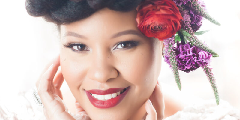 African American woman smiling. Her red lips compliment the red and purple floral crown that adorns her head.