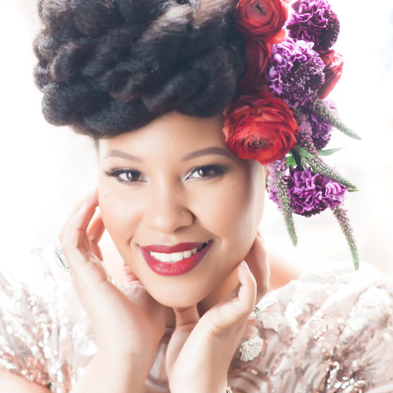 African American woman smiling. Her red lips compliment the red and purple floral crown that adorns her head.